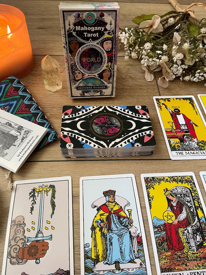 May include: A deck of "Mahogany Tarot" cards with a decorative box featuring diverse faces and the word "WORLD". Several tarot cards are displayed, along with a candle, crystal, and small bouquet. The cards have colorful illustrations.