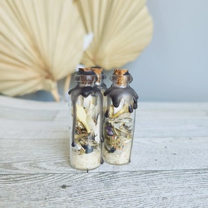 May include: Three small glass bottles with cork stoppers and black wax seals. The bottles are filled with dried herbs and crystals. The bottles are sitting on a light wood surface.