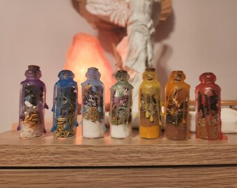 May include: Seven small glass bottles with cork stoppers, each filled with different colored herbs and crystals. The bottles are sealed with colorful wax drippings. The bottles are arranged on a wooden surface.
