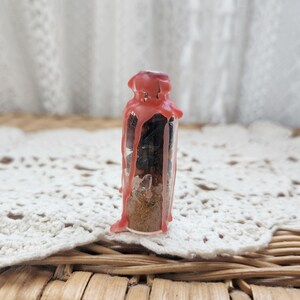 May include: A small, clear glass bottle filled with layers of materials, including a light brown powder and dark, solid pieces. The bottle is sealed with a red wax, which has dripped down the sides. The bottle is set against a white lace background.