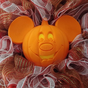 Könnte beinhalten: Ein festlicher Halloween-Kranz mit einem lächelnden orangefarbenen Kürbis mit Micky-Maus-Ohren, umgeben von rotem, weißem und goldenem Netz. Der Kürbis ist 30 cm groß, in einem 36 cm Ring, wodurch ein 46 cm Kranz entsteht. Text: 12" PUMPKIN IN AN 14" RING THAT MAKES AN-18" WREATH WHEN MATERIAL IS ADDED.