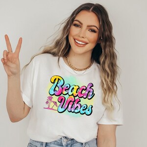 Pu&ograve; includere: T-shirt bianco con la scritta "Beach Vibes" in un font retr&ograve; colorato. Il design include una palma e un ombrellone. La maglietta &egrave; indossata da una persona che fa il segno della pace.