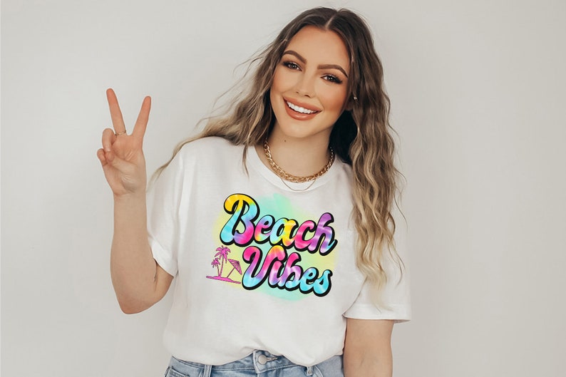 Pu&ograve; includere: T-shirt bianco con la scritta "Beach Vibes" in un font retr&ograve; colorato. Il design include una palma e un ombrellone. La maglietta &egrave; indossata da una persona che fa il segno della pace.