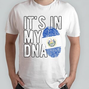 Puede incluir: Camiseta blanca con el texto "IT'S IN MY DNA" en negro, con un dise&ntilde;o de huella dactilar que presenta la bandera de El Salvador.