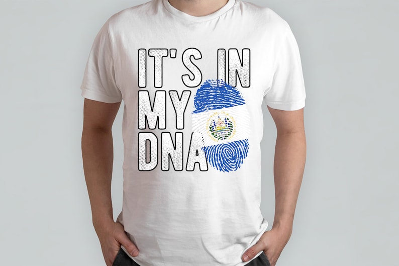 Puede incluir: Camiseta blanca con el texto "IT'S IN MY DNA" en negro, con un dise&ntilde;o de huella dactilar que presenta la bandera de El Salvador.