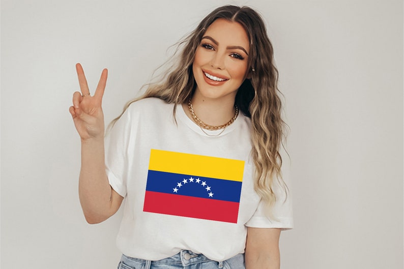 May include: A white t-shirt with a printed image of the Venezuelan flag. The flag is yellow, blue, and red with a star-shaped arc in the center.