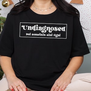 May include: Black t-shirt with white text that reads "Undiagnosed but somethin ain't right".