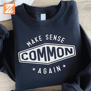 Può includere: Una felpa blu navy con la scritta bianca "MAKE SENSE COMMON AGAIN". La parola "COMMON" è all'interno di una forma geometrica bianca. Due stelle bianche sono sotto la parola "AGAIN".