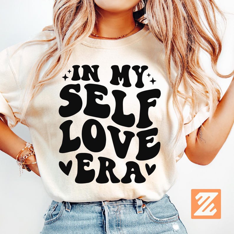 May include: Cream-colored t-shirt with the text "IN MY SELF LOVE ERA" in bold, black, retro-style font. The design includes star accents and heart symbols. The shirt is worn by a person with long, wavy blonde hair.