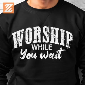 Peut inclure: Sweat-shirt noir avec le texte blanc "WORSHIP WHILE You wait" dans une police vieillie. Le sweat-shirt est de couleur noire unie et présente un design simple et classique.