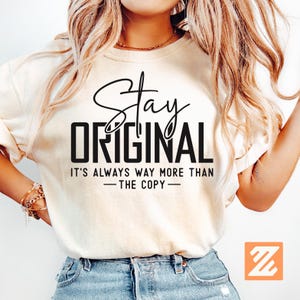 Puede incluir: Camiseta color crema con las palabras "Stay Original" en escritura negra y en negrita. El texto "It's always way more than - the copy -" est&aacute; debajo. La camiseta la lleva una persona con pelo largo y rubio ondulado.