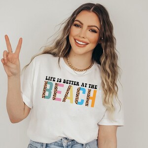 Pu&ograve; includere: T-shirt bianca con la scritta "LIFE IS BETTER AT THE BEACH" in un font colorato e giocoso. La parola "BEACH" presenta un design con stampa leopardata. La maglietta &egrave; un capo d'abbigliamento casual a tema spiaggia.
