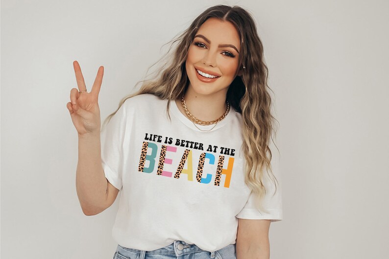 Pu&ograve; includere: T-shirt bianca con la scritta "LIFE IS BETTER AT THE BEACH" in un font colorato e giocoso. La parola "BEACH" presenta un design con stampa leopardata. La maglietta &egrave; un capo d'abbigliamento casual a tema spiaggia.