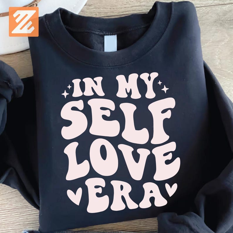 May include: Black crewneck sweatshirt with the text "IN MY SELF LOVE ERA" in a retro font. The text is in a light pink color. The sweatshirt is made of a soft material.