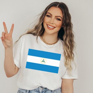 May include: A white t-shirt with a blue and white Nicaraguan flag printed on the front. The flag features a triangle with a green tree and a gold border.