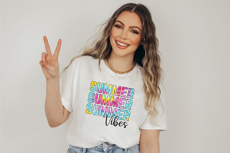 Pu&ograve; includere: T-shirt bianca con la scritta "SUMMER" impilata tre volte in un design tie-dye colorato, con "Vibes" in corsivo sotto. La maglietta &egrave; indossata.