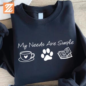 May include: A navy blue sweatshirt with the text "My Needs Are Simple" in white script. Below the text are white illustrations of a coffee cup, a paw print, and an open book. The sweatshirt is made of a soft material.