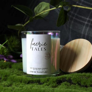 May include: A soy blend wax candle in an iridescent glass jar with a light wood lid. The white label features the text "faerie TALES" and "TWIN THISTLES" with "NET WT 8 OZ". The candle is filled with white wax and displays a circular stamp.