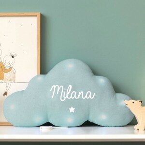 Può includere: Un cuscino a forma di nuvola di colore azzurro chiaro con una stella bianca e il nome "Milana" ricamato. Il cuscino è dotato di luci LED integrate e viene fornito con un telecomando.