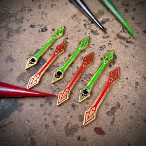 Può includere: Sette spille dorate a forma di freccia con accenti in smalto rosso e verde. Le frecce hanno un design a forma di teschio al centro.