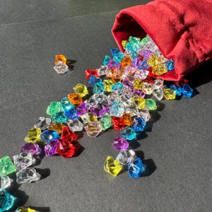 May include: A red velvet bag spills out a variety of colorful, faceted, gem-like stones. The stones are scattered on a black surface.