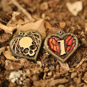 Op de afbeelding: Twee hartvormige pinnen in goudkleur. Eén pin heeft een schedel en gekruiste beenderen ontwerp met rode accenten. De andere pin heeft een rood hart met een gouden nummer "1" in het midden.