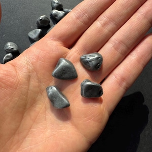 Op de afbeelding: Vijf gladde, grijze stenen met een metallic glans. De stenen zijn van verschillende grootte en vorm en liggen in een hand.