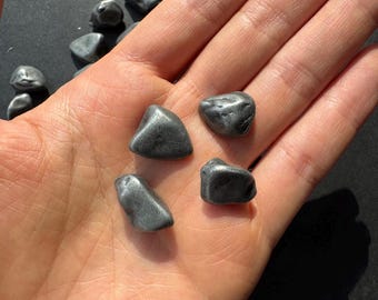 May include: Five smooth, gray stones with a metallic sheen. The stones are various sizes and shapes, and they are arranged in a hand.