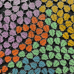 Könnte beinhalten: Eine Sammlung von bunten Metall-Blatt-Pins in verschiedenen Schattierungen von Lila, Orange, Grün und Blau. Die Pins sind in einem Muster angeordnet, das einen Farbverlaufseffekt erzeugt.