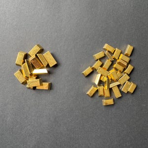 May include: Two piles of small, gold-coloured metal bars. The bars are arranged in a random pattern. The bars are all the same size and shape.