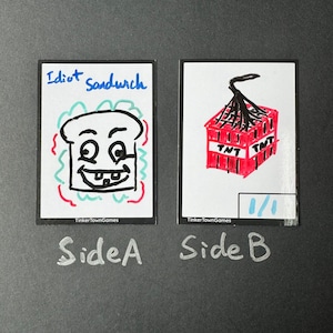 May include: Two white cards with black marker drawings. The card on the left has a cartoon drawing of a white bread slice with a face and the words "Idiot Sandwich" above it. The card on the right has a red drawing of a box of dynamite with a lit fuse and the words "TNT" on the sides. The card on the right also has the number "1/1" written in blue marker.