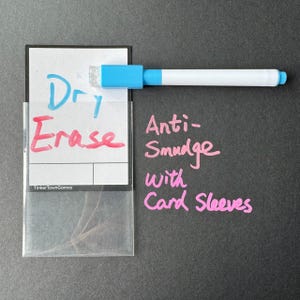 May include: A white dry erase card with a clear plastic sleeve. The card has the words "Dry Erase" written on it in blue and red ink. The text "TinkerTownGames" is printed in black at the bottom of the card. A blue dry erase marker is lying on top of the card. The text "Anti-Smudge with Card Sleeves" is written in pink on a black background.