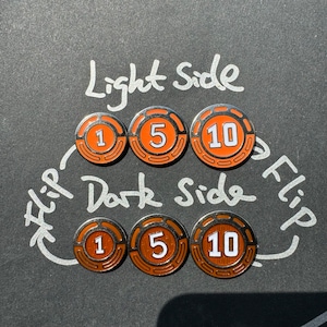 Peut inclure: Six jetons orange et argent avec les chiffres 1, 5 et 10 dessus. Les jetons sont disposés en deux rangées de trois, les chiffres face vers le haut. Le texte "Light Side" est écrit au-dessus des jetons, et le texte "Flip Dark Side Flip" est écrit en dessous des jetons.