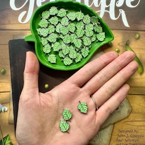 Könnte beinhalten: Eine grüne Blatt-Emaille-Anstecknadel in einer grünen, blattartigen Schale. Vier weitere Nadeln befinden sich auf einer Hand und zeigen die Größe der Nadeln.