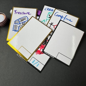 May include: Five blank white cards with gold and black borders. The cards have handwritten words on them, including "Treasure", "Campfire", and "Creeper". The cards are from Tinkertown Games.