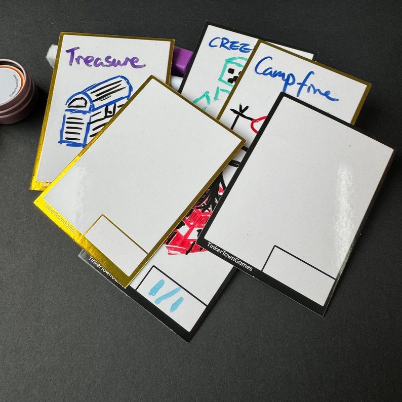 May include: Five blank white cards with gold and black borders. The cards have handwritten words on them, including "Treasure", "Campfire", and "Creeper". The cards are from Tinkertown Games.