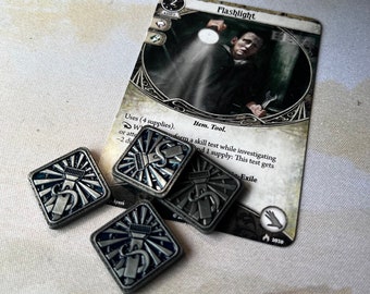Pode incluir: Quatro fichas de prata com um design de lanterna. As fichas estão sobre uma superfície branca ao lado de um cartão de jogo de cartas com o texto "Flashlight" e "Item. Tool." O cartão é do jogo de cartas Arkham Horror: The Card Game.