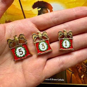 May include: Three gold-coloured metal pins with red, white and green enamel. Each pin features a golden eagle perched atop a red banner with a white circle containing a number. The numbers are 1, 3 and 5.