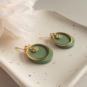 May include: A pair of green and gold earrings with a small flower charm. The earrings are on a white speckled surface.