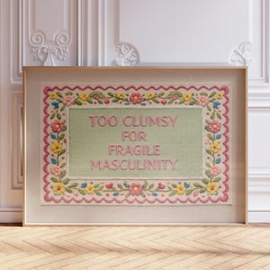 Peut inclure: Une œuvre d'art encadrée au point de croix avec le texte "TOO CLUMSY FOR FRAGILE MASCULINITY". La broderie présente un fond vert clair avec des accents floraux roses, jaunes et bleus. Le cadre est de couleur bois clair.