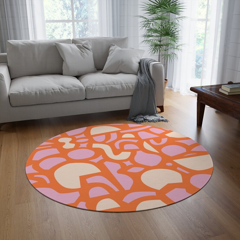 Op de afbeelding: Een rond vloerkleed met een abstract patroon van roze, oranje en cr&egrave;mekleurige vormen. Het vloerkleed ligt op een houten vloer in een woonkamer.