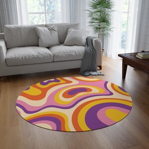 May include: A round rug with a retro design. The rug features a swirling pattern in shades of orange, yellow, purple, and pink, set against a cream background. The rug is placed on a hardwood floor in a living room setting.