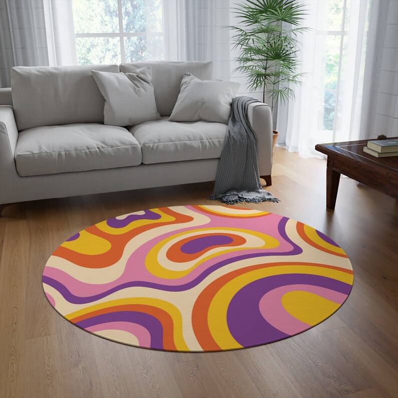 May include: A round rug with a retro design. The rug features a swirling pattern in shades of orange, yellow, purple, and pink, set against a cream background. The rug is placed on a hardwood floor in a living room setting.