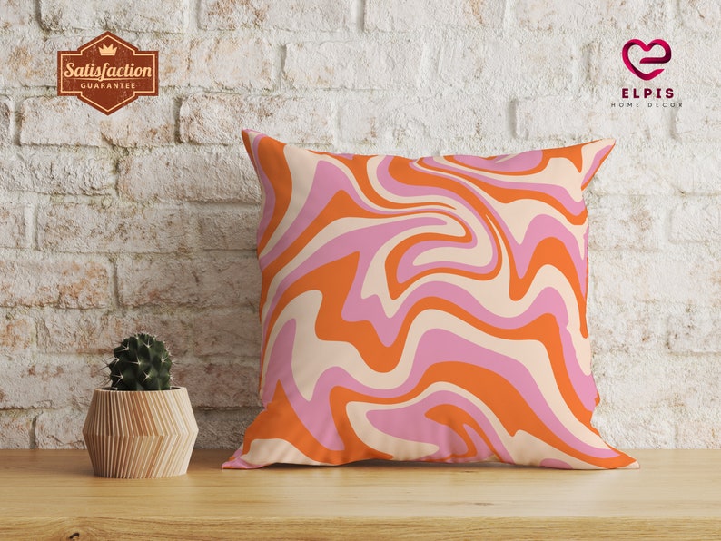 Pode incluir: Travesseiro de decora&ccedil;&atilde;o com padr&atilde;o de redemoinhos laranja e rosa sobre fundo branco. O travesseiro est&aacute; colocado sobre uma superf&iacute;cie de madeira em frente a uma parede de tijolo branco.