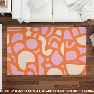 Op de afbeelding: Abstract vloerkleed met een geometrisch patroon in oranje, roze en cr&egrave;me. Het vloerkleed wordt getoond op een houten vloer met een witte bank en een pluche wit vloerkleed op de achtergrond.
