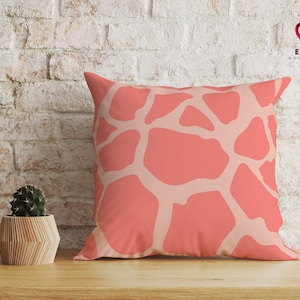 May include: A coral pink throw pillow with a white giraffe print pattern. The pillow is on a wooden surface in front of a white brick wall.