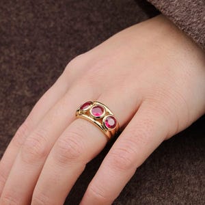 Op de afbeelding: Een gouden ring met drie ovale rode edelstenen. De ring is gezet in een gouden band en wordt aan een hand gedragen. De achtergrond omvat een bruine jas en doorschijnende panty's.