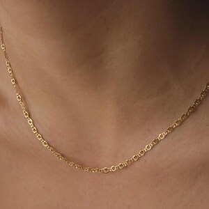 May include: A delicate gold chain necklace featuring a repeating pattern of small, circular links connected by thin, straight bars. The necklace rests against the skin, showcasing a polished, metallic finish that reflects light across its surface.