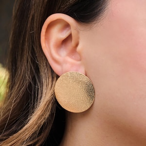 May include: A large, gold-colored, textured disc earring is shown. The earring is round and has a slightly hammered appearance. The earring is worn on the ear of a person with brown hair.