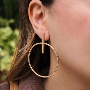 May include: Gold-toned hoop earrings with a vertical bar accent. The earrings have a minimalist design, featuring a large circular hoop and a straight, rectangular bar that intersects the hoop. The jewelry is shown on a person.